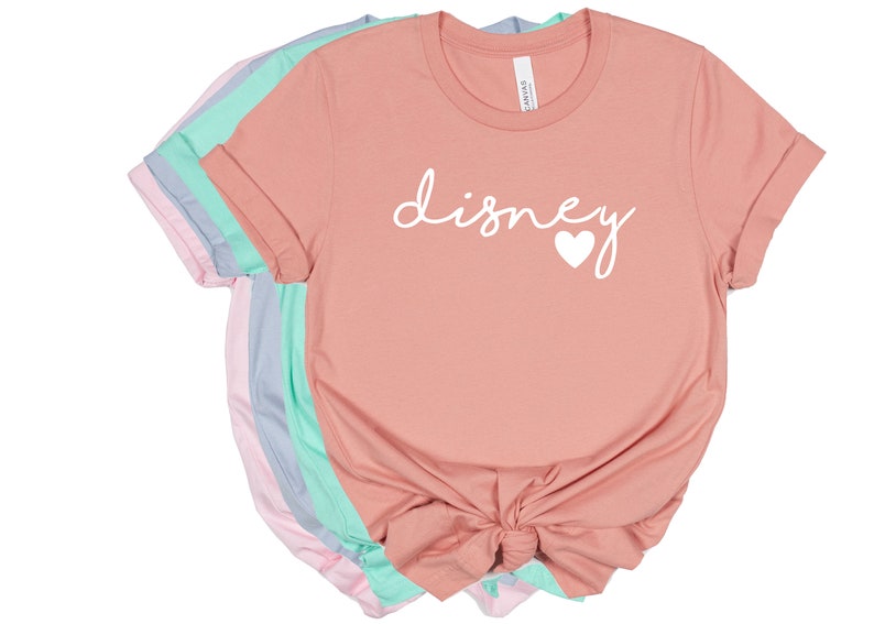 May include: A stack of t-shirts in various pastel colors, including pink, blue, and mint green. The front shirt is peach-colored and features the word "disney" in white script with a white heart. The shirts are folded with rolled sleeves.