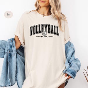 May include: A cream-colored t-shirt with the text "Volleyball Mom" in black lettering. A black and white volleyball graphic is included in the text.