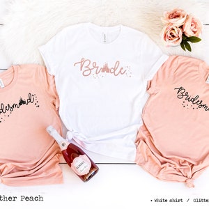 Può includere: Tre magliette bianche con testo nero e un design di castello rosa glitterato. Le magliette dicono "Bride" e "Bridesmaid". Le magliette sono su uno sfondo di legno bianco con una bottiglia di champagne rosa e rose rosa.
