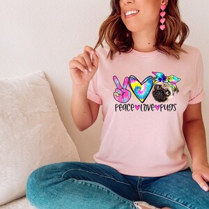 May include: A pink t-shirt with a tie-dye peace sign, heart, and pug design. The text "Peace Love Pugs" is printed below the design.