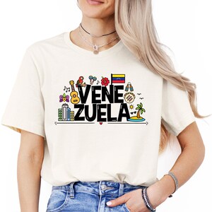 May include: A white t-shirt with a black graphic design that says "VENEZUELA" with colorful illustrations of Venezuelan culture, including a parrot, a guitar, a flag, and a palm tree.