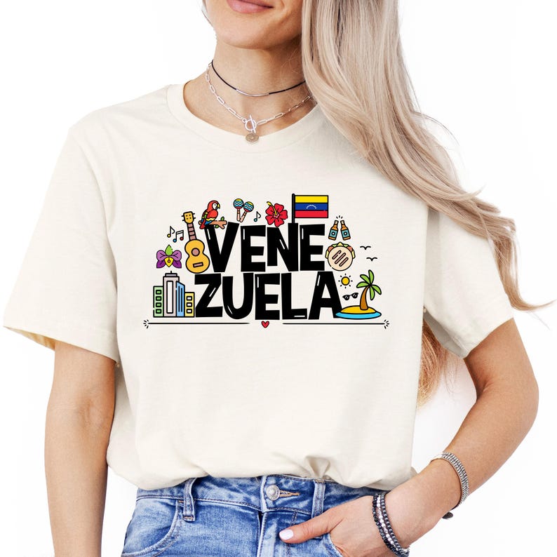 May include: A white t-shirt with a black graphic design that says "VENEZUELA" with colorful illustrations of Venezuelan culture, including a parrot, a guitar, a flag, and a palm tree.