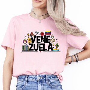 May include: Pink t-shirt with a black graphic design that says "Venezuela" with colorful icons representing the country's culture.