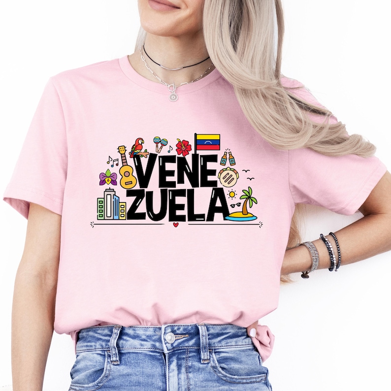 May include: Pink t-shirt with a black graphic design that says "Venezuela" with colorful icons representing the country's culture.
