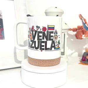 May include: White ceramic travel mug with a cork lid. The mug features a colorful graphic design with the word "VENEZUELA" in black letters and various icons representing the country, including a guitar, a palm tree, a flag, and a hibiscus flower.
