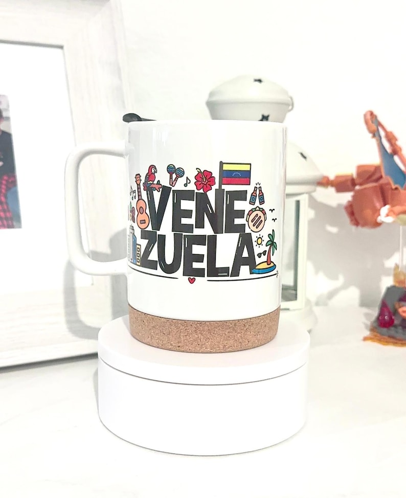 May include: White ceramic travel mug with a cork lid. The mug features a colorful graphic design with the word "VENEZUELA" in black letters and various icons representing the country, including a guitar, a palm tree, a flag, and a hibiscus flower.