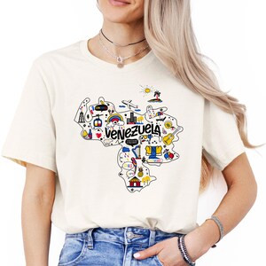 Może przedstawiać: Biały t-shirt z kolorową grafiką Wenezueli. Grafika zawiera mapę kraju z r&oacute;żnymi ikonami reprezentującymi zabytki, kulturę i przyrodę. Tekst "Venezuela" jest napisany pogrubioną, kolorową czcionką.