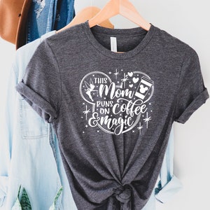 May include: A gray t-shirt with a white graphic design that says "This Mom Runs on Coffee & Magic". The graphic design features a Mickey Mouse ear silhouette with a coffee cup, a fairy, and hearts.
