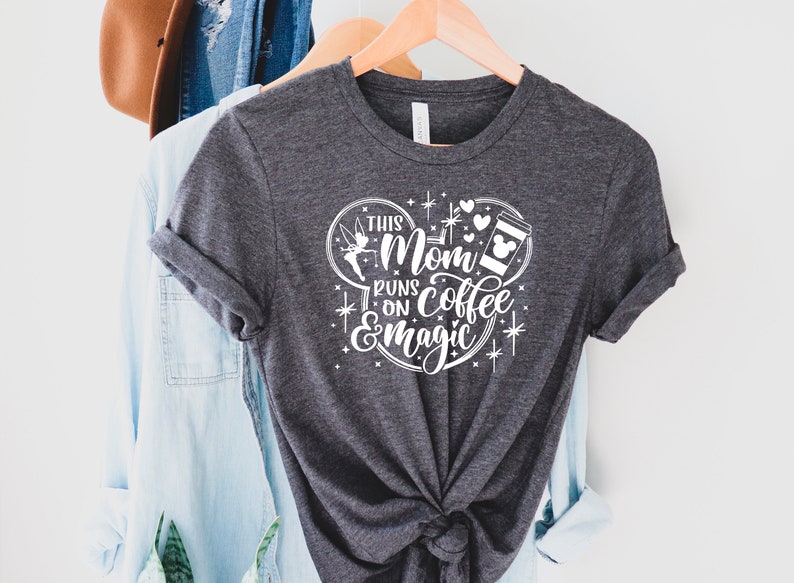 May include: A gray t-shirt with a white graphic design that says "This Mom Runs on Coffee & Magic". The graphic design features a Mickey Mouse ear silhouette with a coffee cup, a fairy, and hearts.