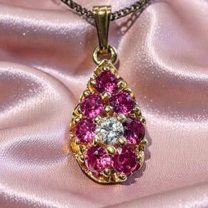 Puede incluir: Un collar con colgante de oro con un diseño en forma de lágrima. El colgante presenta un grupo de piedras preciosas rosas que rodean una piedra preciosa central transparente. La cadena del collar es de color marrón oscuro, sobre un fondo de satén rosa.
