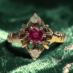 Op de afbeelding: Een gouden ring met een centrale, ronde, dieprode edelsteen, geflankeerd door twee kleinere, heldere edelstenen. De ring heeft een sierlijk filigraan ontwerp, tegen een groene achtergrond.