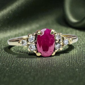May include: A gold ring featuring a large oval ruby center stone, surrounded by smaller clear gemstones. The ring has a classic design with a polished gold band and is set against a green background.