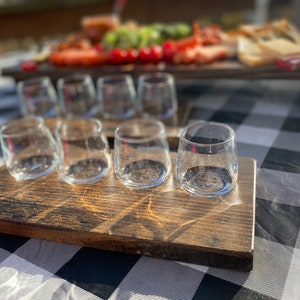 May include: A wooden serving tray with six clear glass shot glasses. The tray is dark brown and has a rustic finish. The glasses are arranged in a row on the tray.