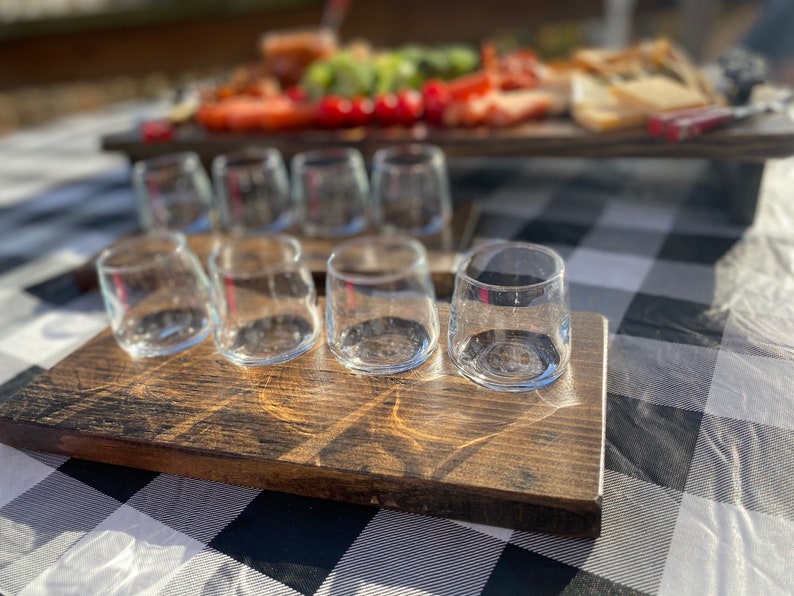 May include: A wooden serving tray with six clear glass shot glasses. The tray is dark brown and has a rustic finish. The glasses are arranged in a row on the tray.