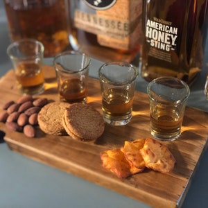 May include: A wooden serving tray with four shot glasses filled with amber liquid, two shortbread cookies, almonds, and a few crispy crackers. The tray is next to three bottles of whiskey, one labeled "Wild Turkey American Honey Sting".