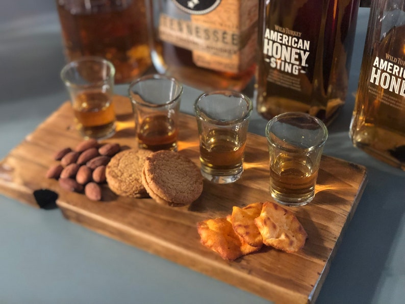 May include: A wooden serving tray with four shot glasses filled with amber liquid, two shortbread cookies, almonds, and a few crispy crackers. The tray is next to three bottles of whiskey, one labeled "Wild Turkey American Honey Sting".