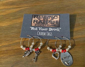 May include: Set of three wine glass charms with red beads and silver charms. The charms are a key, a heart, and an oval with the word "always" on it. The card reads "Rustic Ranch Creations" and "Not Your Drink" Charm Tags.