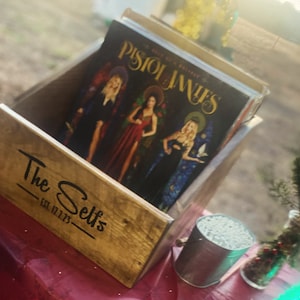 May include: A wooden crate with the text "The Selts Est. 12.12.73" on the front. The crate is filled with vinyl records, including one with the title "Pistol Janes - Bells of a Broken Trinity".