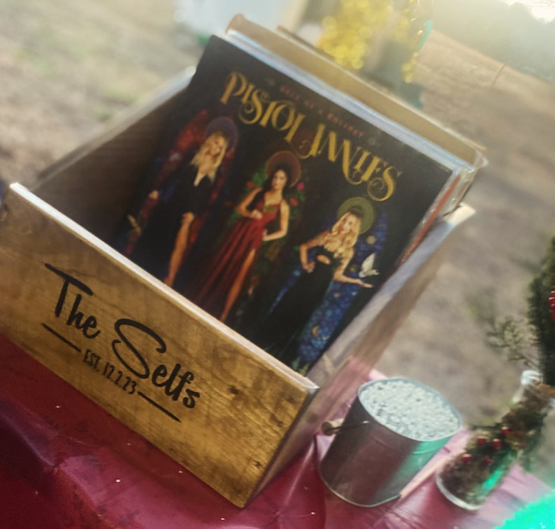 May include: A wooden crate with the text "The Selts Est. 12.12.73" on the front. The crate is filled with vinyl records, including one with the title "Pistol Janes - Bells of a Broken Trinity".