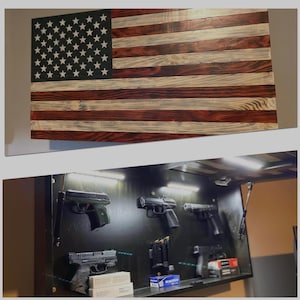 Puede incluir: Una bandera americana de madera pintada en rojo, blanco y azul, colgada sobre una caja fuerte de armas negra con un estante y varias armas de fuego y municiones.