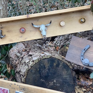 Puede incluir: Estante de madera rústica con un cráneo de vaca, ganchos de herradura y un gancho para botas. El estante está montado en un tronco de árbol.