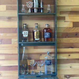 Op de afbeelding: Een houten bar-kast met een neerklappende voorkant. De kast heeft twee planken met flessen drank, glazen en een cocktailshaker. De kast is donkergroen geverfd en heeft een rustieke uitstraling. De kast staat op een houten aanrecht met twee mokken waarop "Christmas Cheer" en "Christmas Trees" staat.