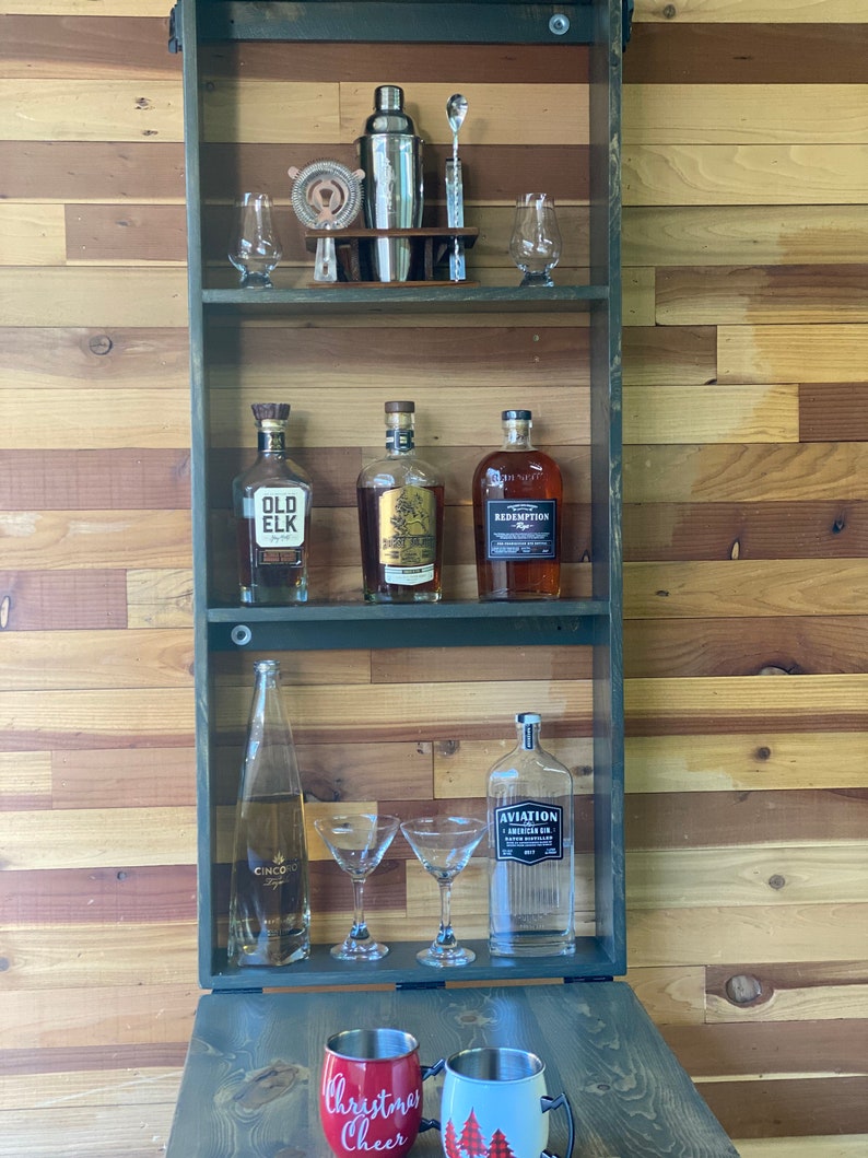 Op de afbeelding: Een houten bar-kast met een neerklappende voorkant. De kast heeft twee planken met flessen drank, glazen en een cocktailshaker. De kast is donkergroen geverfd en heeft een rustieke uitstraling. De kast staat op een houten aanrecht met twee mokken waarop "Christmas Cheer" en "Christmas Trees" staat.