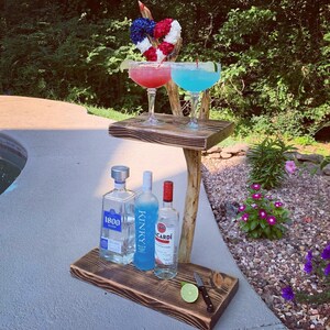 May include: A wooden two-tiered serving tray with a natural branch design. The tray is holding two margarita glasses, one with a blue drink and one with a pink drink. There are three bottles of liquor on the lower shelf: 1800 Tequila, Kinky Liqueur, and Bacardi Rum. A lime wedge and a small knife are also on the lower shelf.