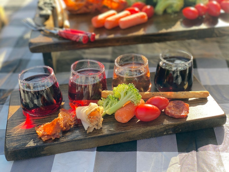 May include: A wooden serving board with four small glasses filled with red wine, a small plate of sliced meat, a few pieces of bacon, a small bunch of broccoli, a few carrot sticks, and a few cherry tomatoes.