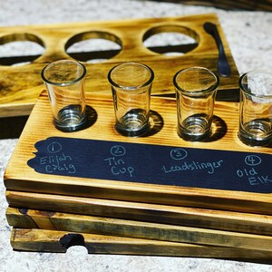 May include: Wooden tray with four shot glasses and a chalkboard label with the names of four different drinks: Elijah Craig, Tin Cup, Lead Slinger, and Old Elk.