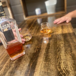 Op de afbeelding: Een doorzichtige glazen fles met een bruine vloeistof en een zwart etiket met de tekst "Heaven's Door" op een houten tafel met twee doorzichtige glazen gevuld met een bruine vloeistof.