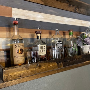 May include: A wooden shelf with a rustic finish, mounted on a wall with a wood plank design. The shelf holds four bottles of liquor, including Tito's Handmade Vodka, Old Elk Bourbon, and Aviation American Gin. There are also four clear glass shot glasses on the shelf.