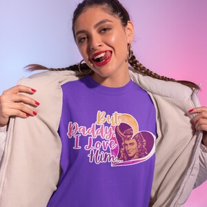 May include: A purple t-shirt featuring a graphic of a heart shape containing an illustration of a long-haired character with horns. The text "But Daddy, I Love Him..." is printed in a stylized pink and orange font next to the heart graphic.