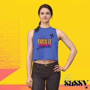 May include: Blue crop top with the text "FUCK IT BOOK IT" in orange, pink, and purple. The design includes a black airplane and a dotted line with a location pin. The background is yellow.