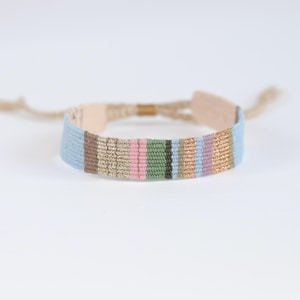Op de afbeelding: Een veelkleurige geweven armband met lichtblauwe, roze, bruine, gouden en groene strepen. De armband is met een knoop vastgemaakt en heeft een kleine gouden kraal.