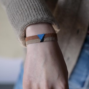 May include: A woven bracelet with a brown, tan, and blue geometric pattern. The bracelet is made of thin, braided cords.