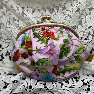 Könnte beinhalten: Eine kleine, rosa Münzgeldbörse mit Metallverschluss und Blumen- und Marienkäfermuster. Die Geldbörse hat einen Kiss-Lock-Verschluss und ein farbenfrohes Design aus Blumen, Marienkäfern und Blättern.