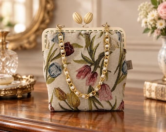 Peut inclure: Un sac à main rectangulaire en tissu orné d'un motif floral tissé de tulipes rouges, jaunes et bleues sur fond beige. Le sac possède un cadre crème avec un fermoir à double bouton et une chaîne dorée agrémentée de perles blanches.