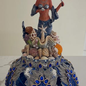 Könnte beinhalten: Eine dekorative Meerjungfrauenfigur mit blauem Schwanz und Krone, die eine Perle hält, platziert auf einem Sockel, der mit verschiedenen Muscheln verziert ist. Der Sockel ist eine abgerundete, hohle Struktur mit silberner Spitze und blauen tropfenförmigen Edelsteinen.