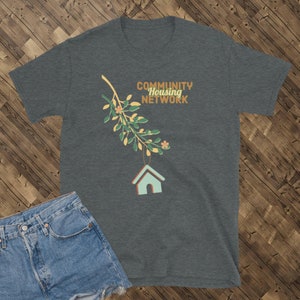 May include: Dark gray t-shirt with the text "COMMUNITY HOUSING NETWORK" in orange and green. A branch with green leaves, small orange flowers, and a hanging teal house graphic is printed on the shirt. Denim shorts are visible in the image.
