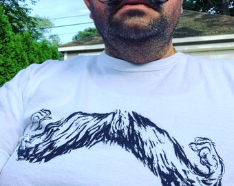 May include: White t-shirt featuring a large, stylized black and white illustration of a mustache. The design has a textured, brushstroke appearance. The shirt is a crew neck style.