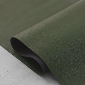 May include: A close-up shot of a textured, olive green material, possibly leather or a similar fabric. The material is rolled, revealing a dark, possibly black, underside. The surface has a subtle, speckled pattern, and the lighting highlights the texture.