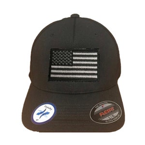 May include: Black baseball cap with a white and grey American flag patch on the front. The cap has a black Flexfit logo on the back with the text "The Original Flexfit L/XL It Fits Your Life".