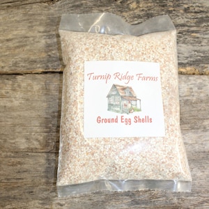 May include: A clear plastic bag filled with ground eggshells. The bag has a white label with the text "Turnip Ridge Farms" and a drawing of a small house. The label also says "Ground Egg Shells".