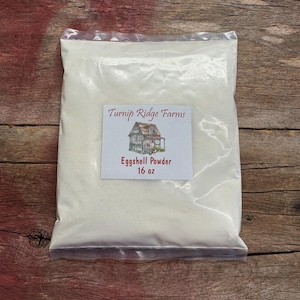 May include: A clear plastic bag filled with white powder, labeled "Turnip Ridge Farms Eggshell Powder 16 oz." The label features a drawing of a small house. The bag is set against a rustic wooden background.