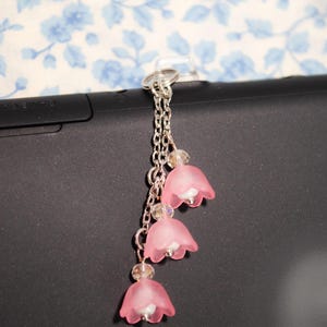 Puede incluir: Un llavero plateado con tres colgantes de flores rosas y detalles de cuentas transparentes. Los pétalos de las flores son translúcidos y cuelgan de una cadena plateada. El fondo tiene un estampado floral.