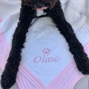May include: A white and pink quilted dog bed with a pink paw print and the name "Olive" embroidered on it. A black dog is lying on the bed.