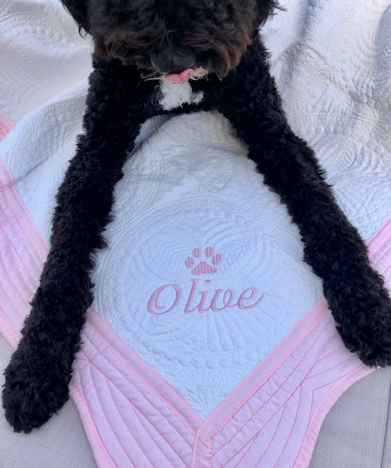 May include: A white and pink quilted dog bed with a pink paw print and the name "Olive" embroidered on it. A black dog is lying on the bed.