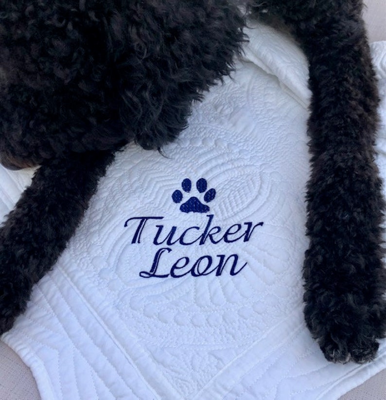 May include: White quilted blanket with a blue paw print and the name "Tucker Leon" embroidered on it. A black dog is lying on the blanket.