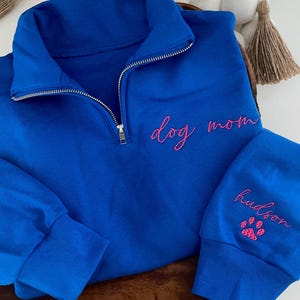 May include: Royal blue pullover sweatshirt with a quarter-zip neckline. The words "dog mom" are embroidered in pink on the chest. The sleeve cuff features the name "hudson" and a pink paw print.